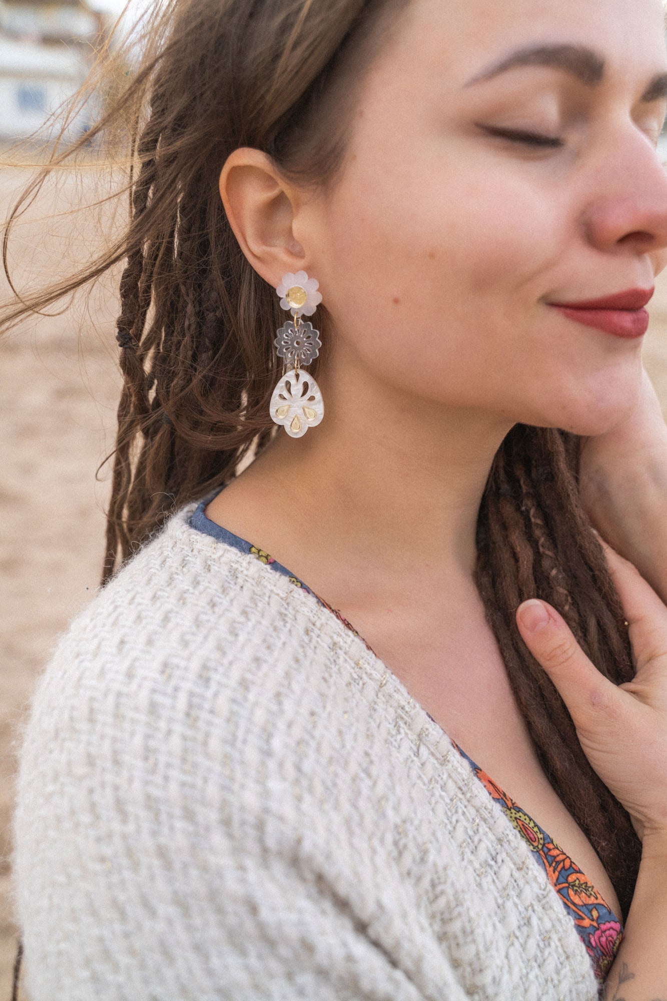 A woman with closed eyes and long brown dreadlocks wears a beige sweater and large handcrafted boho earrings, standing outdoors on a sandy beach. She smiles softly and touches her chest with one hand.