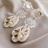 A pair of elegant, handcrafted boho earrings featuring white floral studs, clear flower-shaped connectors, and cream-colored teardrop pendants with gold accents, displayed on soft white fabric with delicate dried grass above.