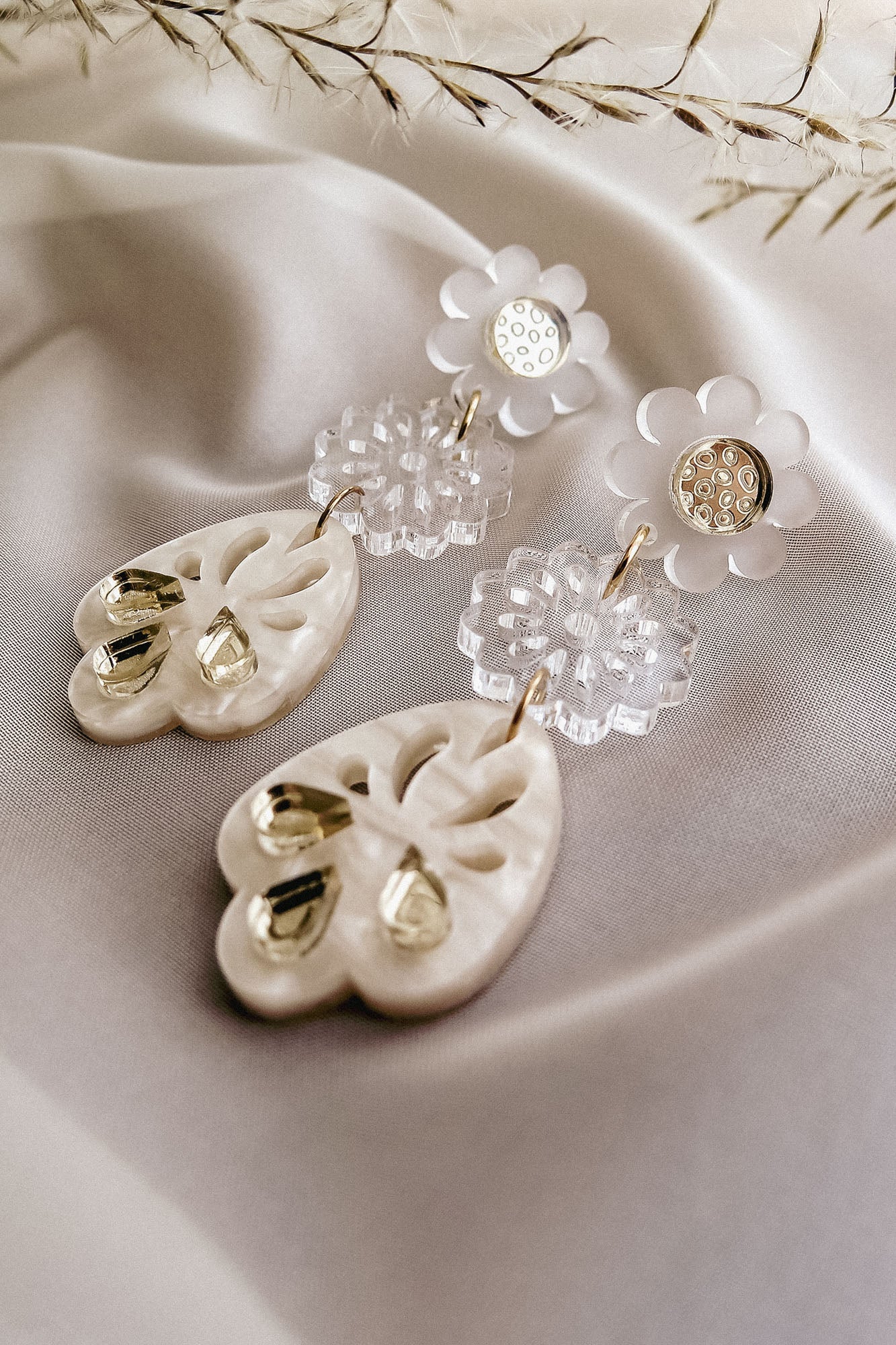 A pair of elegant, handcrafted boho earrings featuring white floral studs, clear flower-shaped connectors, and cream-colored teardrop pendants with gold accents, displayed on soft white fabric with delicate dried grass above.