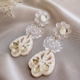A pair of elegant boho earrings featuring white floral studs, clear flower-shaped links, and handcrafted cream-colored, heart-shaped pendants with teardrop accents, resting on a soft, light satin fabric.