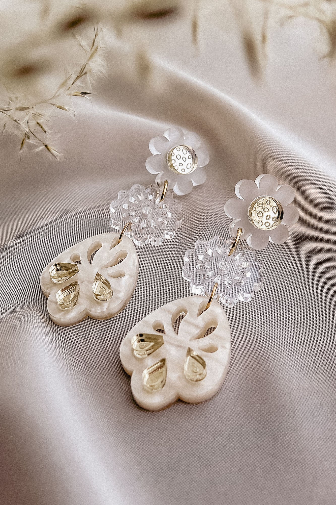 A pair of elegant boho earrings featuring white floral studs, clear flower-shaped links, and handcrafted cream-colored, heart-shaped pendants with teardrop accents, resting on a soft, light satin fabric.
