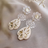 A pair of elegant, handcrafted boho earrings featuring flower-shaped clear studs and ornate, heart-shaped white pendants with gold accents, displayed on soft fabric with delicate dried flowers nearby.