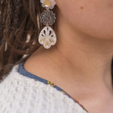 A close-up of a person wearing handcrafted, dangling earrings with flower and abstract shapes in translucent and gold tones, paired with a white textured sweater and a blue shirt with a floral pattern.