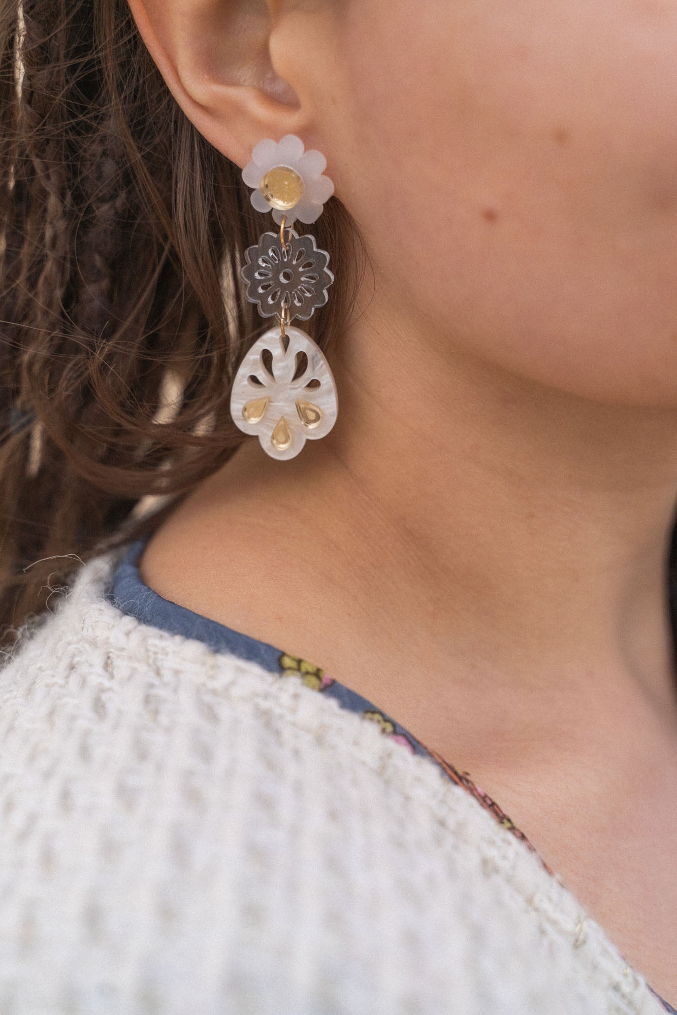 A close-up of a person wearing handcrafted, dangling earrings with flower and abstract shapes in translucent and gold tones, paired with a white textured sweater and a blue shirt with a floral pattern.