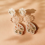 A pair of handcrafted dangling earrings with white floral studs, clear floral middle segments, and translucent, petal-shaped drops featuring cut-out and gold accents, displayed on a peach-colored surface with soft shadows.