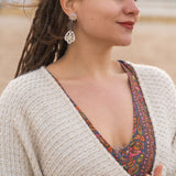 A woman with long braided hair stands outdoors, wearing a patterned dress and a cream knit sweater. She has handcrafted boho earrings and is smiling softly, looking to the side. The background appears to be a beach or sandy area.
