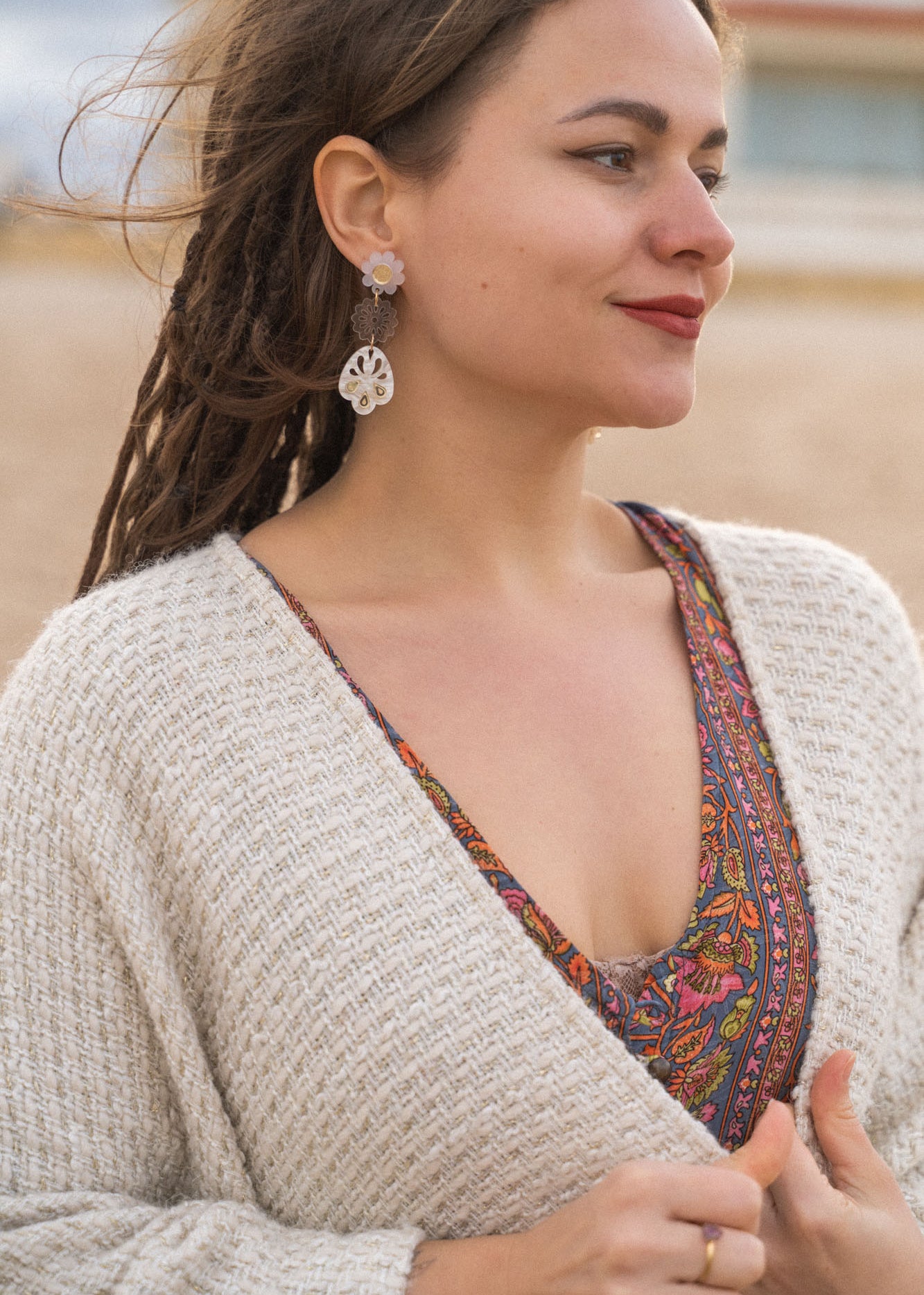 A woman with long braided hair stands outdoors, wearing a patterned dress and a cream knit sweater. She has handcrafted boho earrings and is smiling softly, looking to the side. The background appears to be a beach or sandy area.