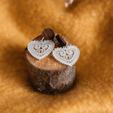 A pair of Folk Heart Earrings with white, lace-patterned hearts and wooden studs embody lightweight elegance, displayed on a small tree stump against golden brown fabric. Delicate dried flowers are visible at the top.