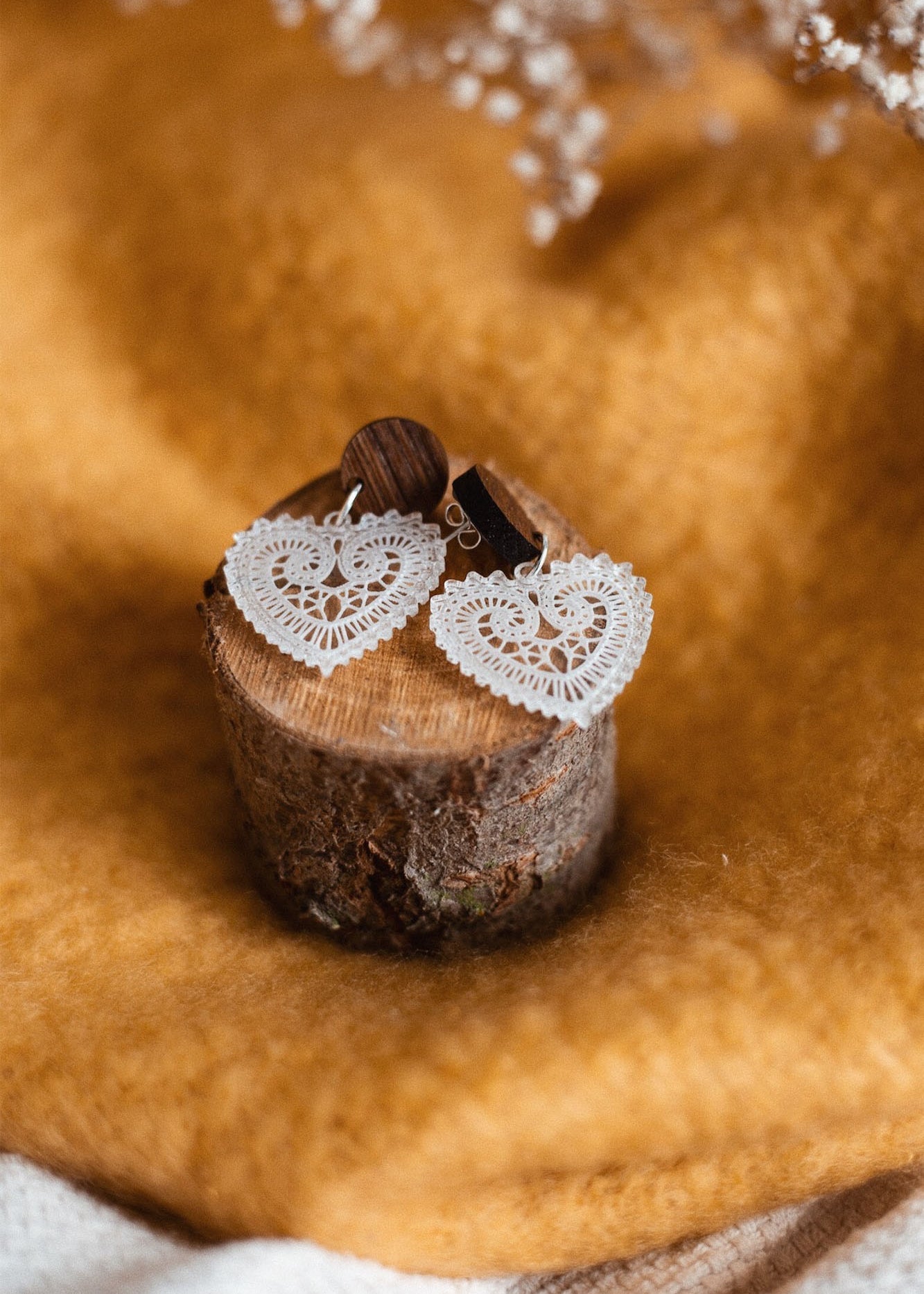 A pair of Folk Heart Earrings with white, lace-patterned hearts and wooden studs embody lightweight elegance, displayed on a small tree stump against golden brown fabric. Delicate dried flowers are visible at the top.
