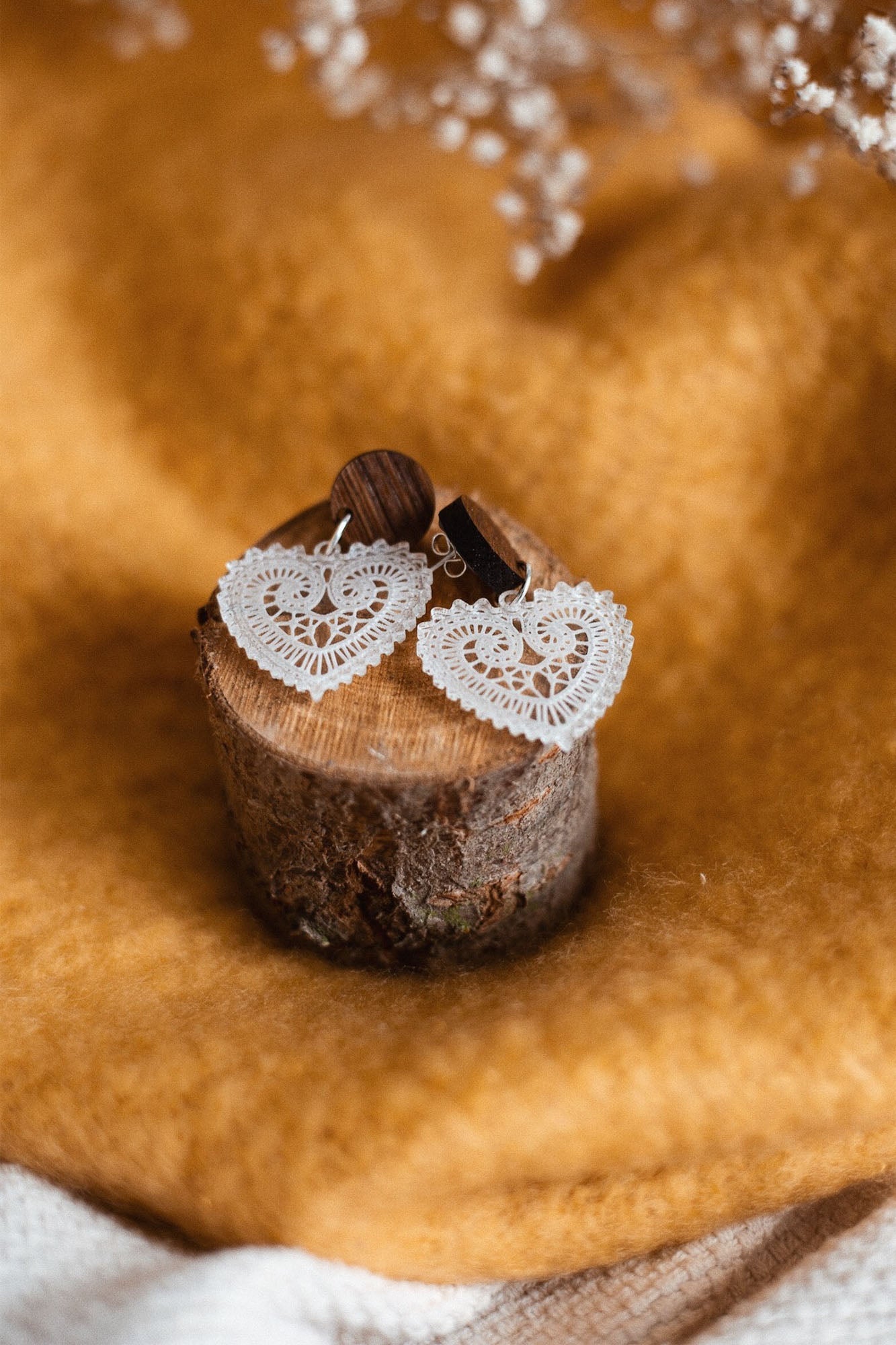 A pair of Folk Heart Earrings with white, lace-patterned hearts and wooden studs embody lightweight elegance, displayed on a small tree stump against golden brown fabric. Delicate dried flowers are visible at the top.