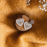 A pair of Folk Heart Earrings, showcasing lightweight elegance and delicate, white lace heart-shaped design, displayed on a small wooden disk atop soft golden-yellow fabric with white babys breath flowers in the background.
