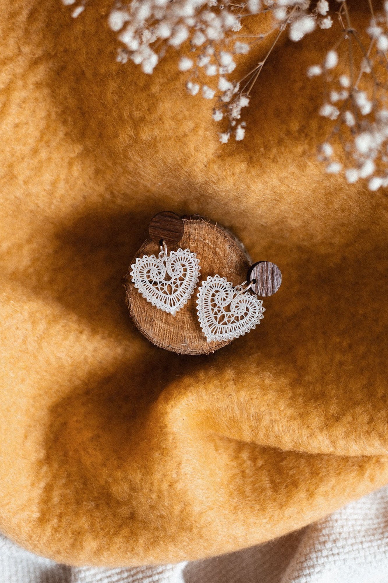 A pair of Folk Heart Earrings, showcasing lightweight elegance and delicate, white lace heart-shaped design, displayed on a small wooden disk atop soft golden-yellow fabric with white babys breath flowers in the background.