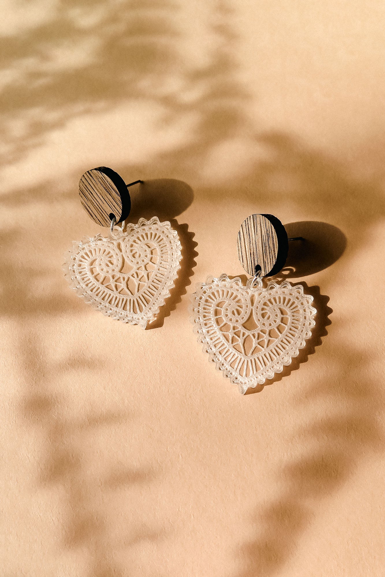 A pair of Folk Heart Earrings with wooden circular studs and intricate, lace-like craftsmanship form lightweight elegance, resting on a beige surface with soft shadows.