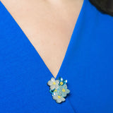 A close-up of a person wearing a vibrant blue V-neck top, adorned with a small, handmade lapel pin featuring blue, yellow, and white forget-me-not flowers pinned near the neckline.