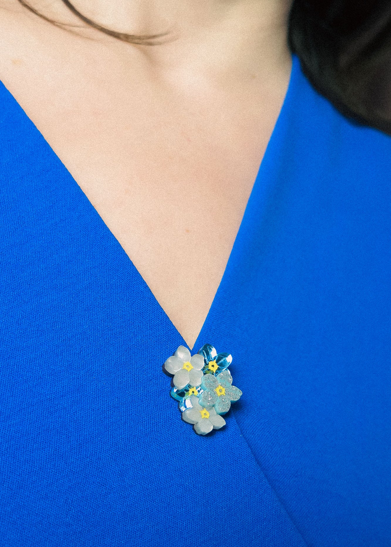 A close-up of a person wearing a vibrant blue V-neck top, adorned with a small, handmade lapel pin featuring blue, yellow, and white forget-me-not flowers pinned near the neckline.