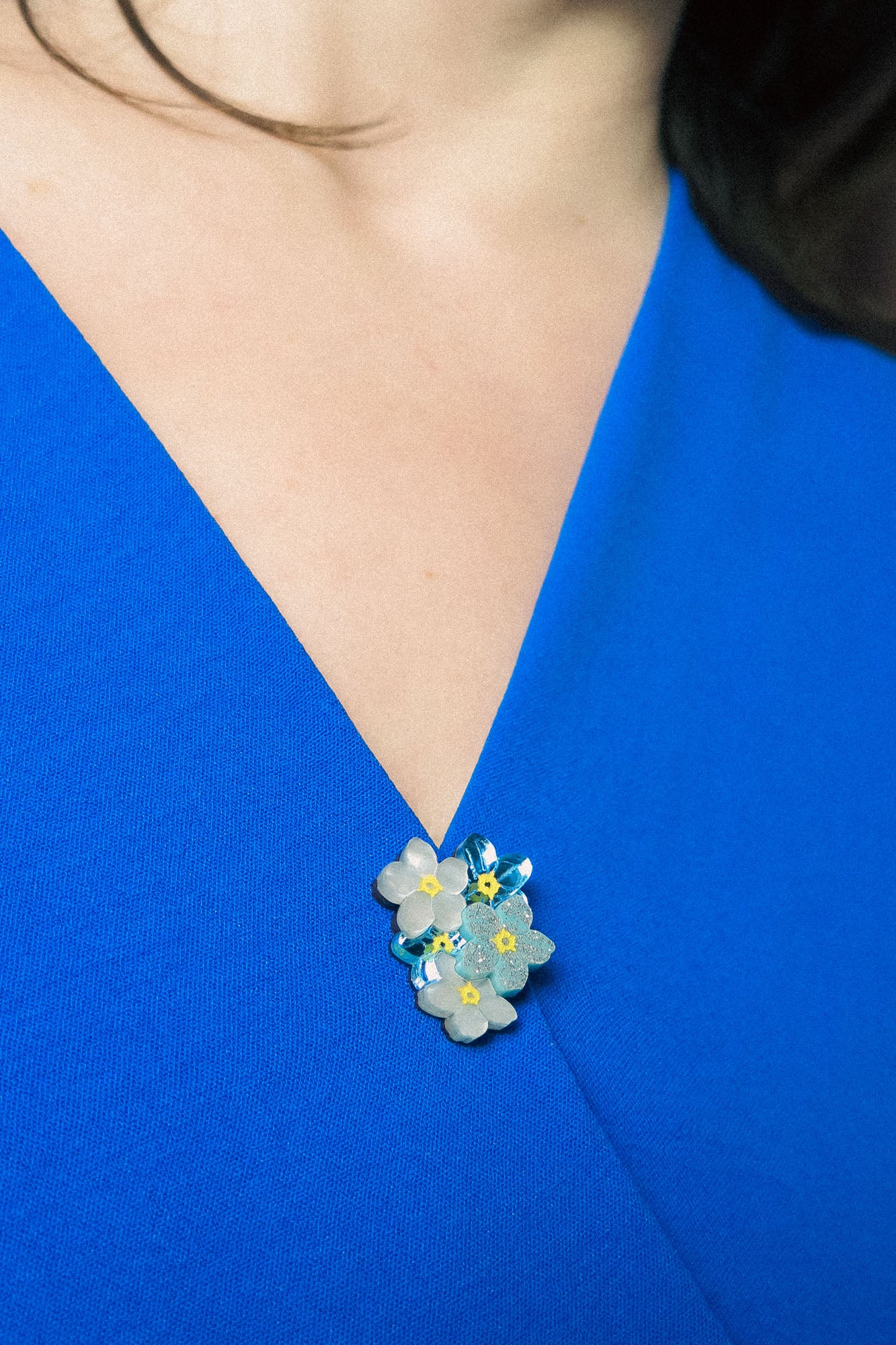 A close-up of a person wearing a vibrant blue V-neck top, adorned with a small, handmade lapel pin featuring blue, yellow, and white forget-me-not flowers pinned near the neckline.