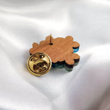 A handmade wooden enamel Lapel Pin shaped like a Forget Me Not Flower with a gold-colored butterfly clasp is displayed face down on smooth white satin fabric, highlighting its detailed back and elegant fastening.