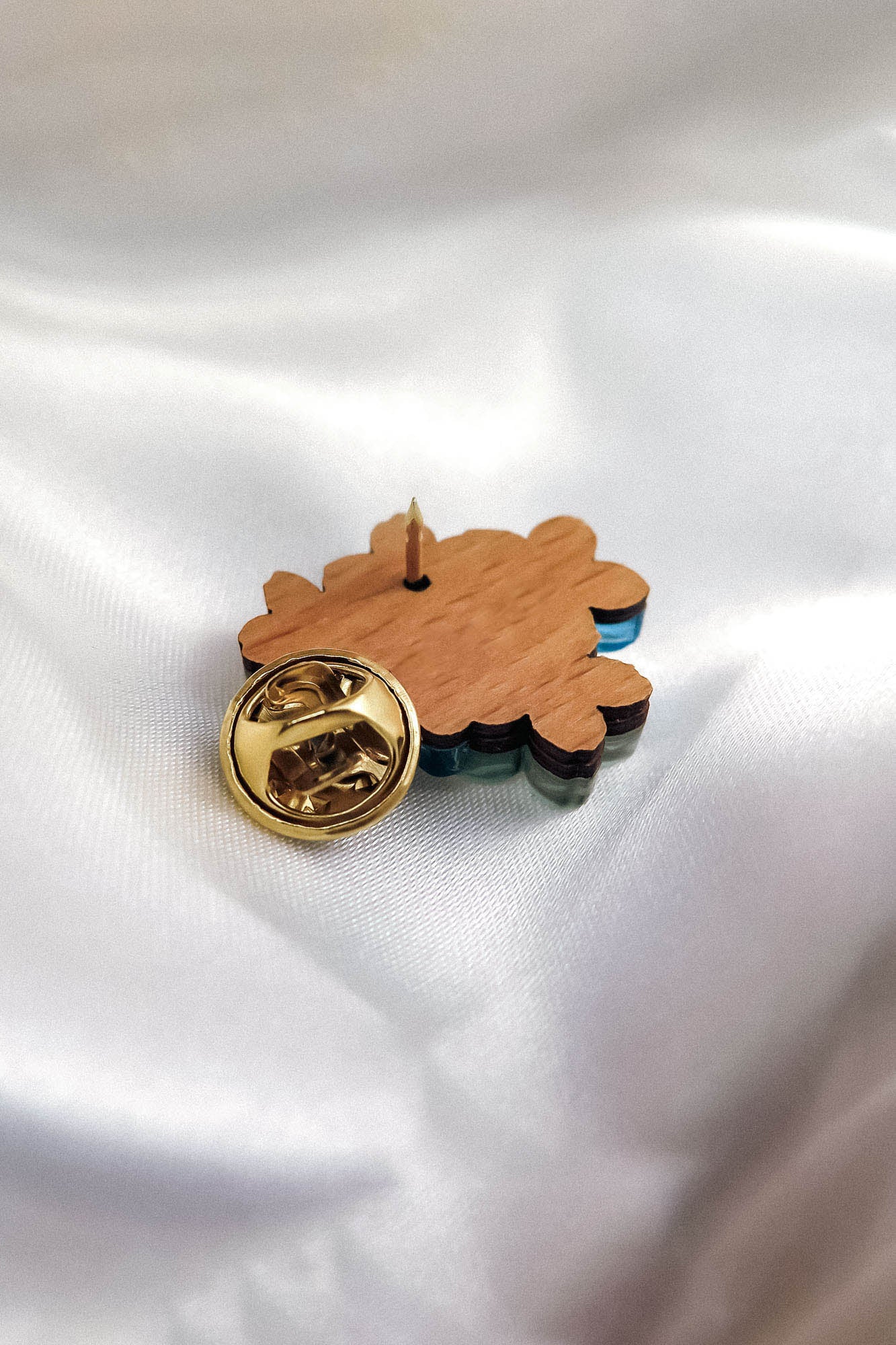 A handmade wooden enamel Lapel Pin shaped like a Forget Me Not Flower with a gold-colored butterfly clasp is displayed face down on smooth white satin fabric, highlighting its detailed back and elegant fastening.