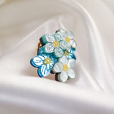 A cluster of handmade pastel blue and white enamel Forget Me Not Flower lapel pins with yellow centers, some accented with glitter, displayed on smooth, white satin fabric.