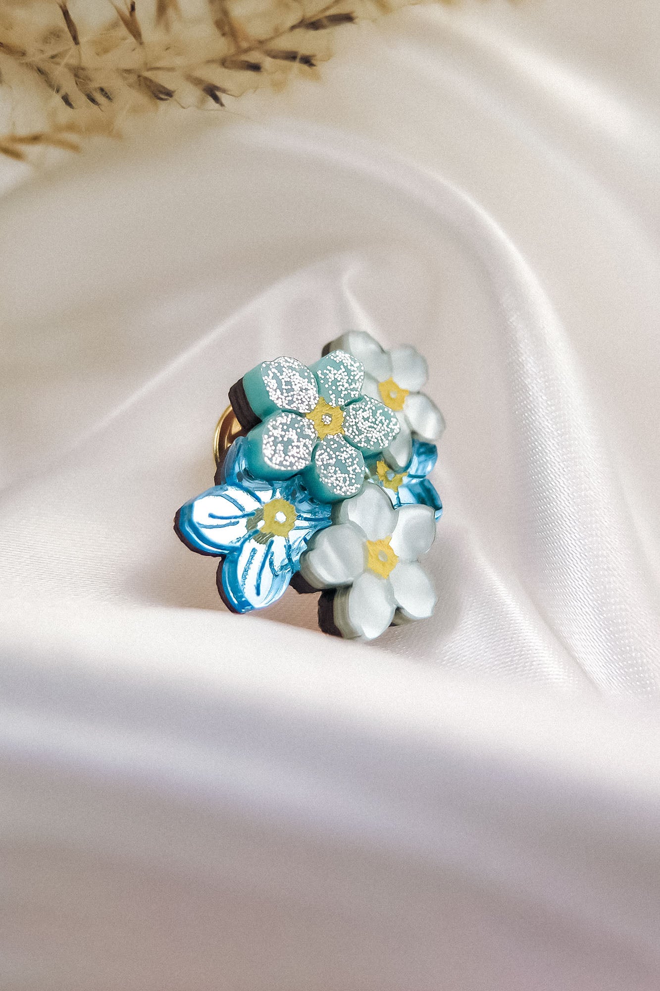 A cluster of handmade pastel blue and white enamel Forget Me Not Flower lapel pins with yellow centers, some accented with glitter, displayed on smooth, white satin fabric.