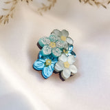 A cluster of handmade flower pins, inspired by the Forget Me Not Flower, in blue and white, rests on soft cream-colored fabric, with delicate dried grass in the background.