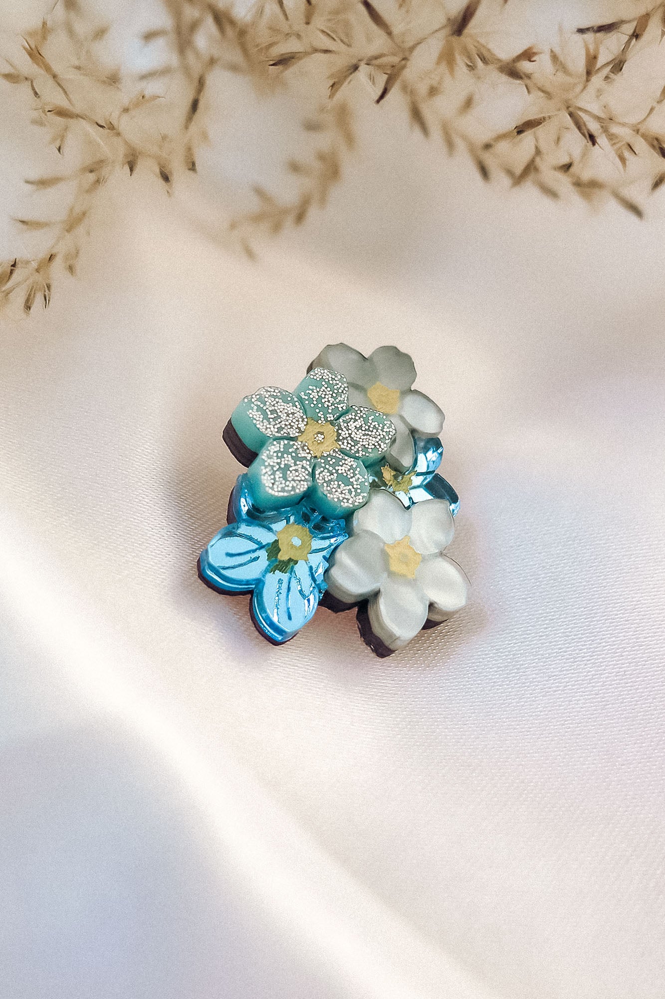 A cluster of handmade flower pins, inspired by the Forget Me Not Flower, in blue and white, rests on soft cream-colored fabric, with delicate dried grass in the background.