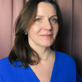 A woman with shoulder-length brown hair wearing a blue top and a handmade Forget Me Not Flower lapel pin stands in front of mauve curtains, smiling gently at the camera.