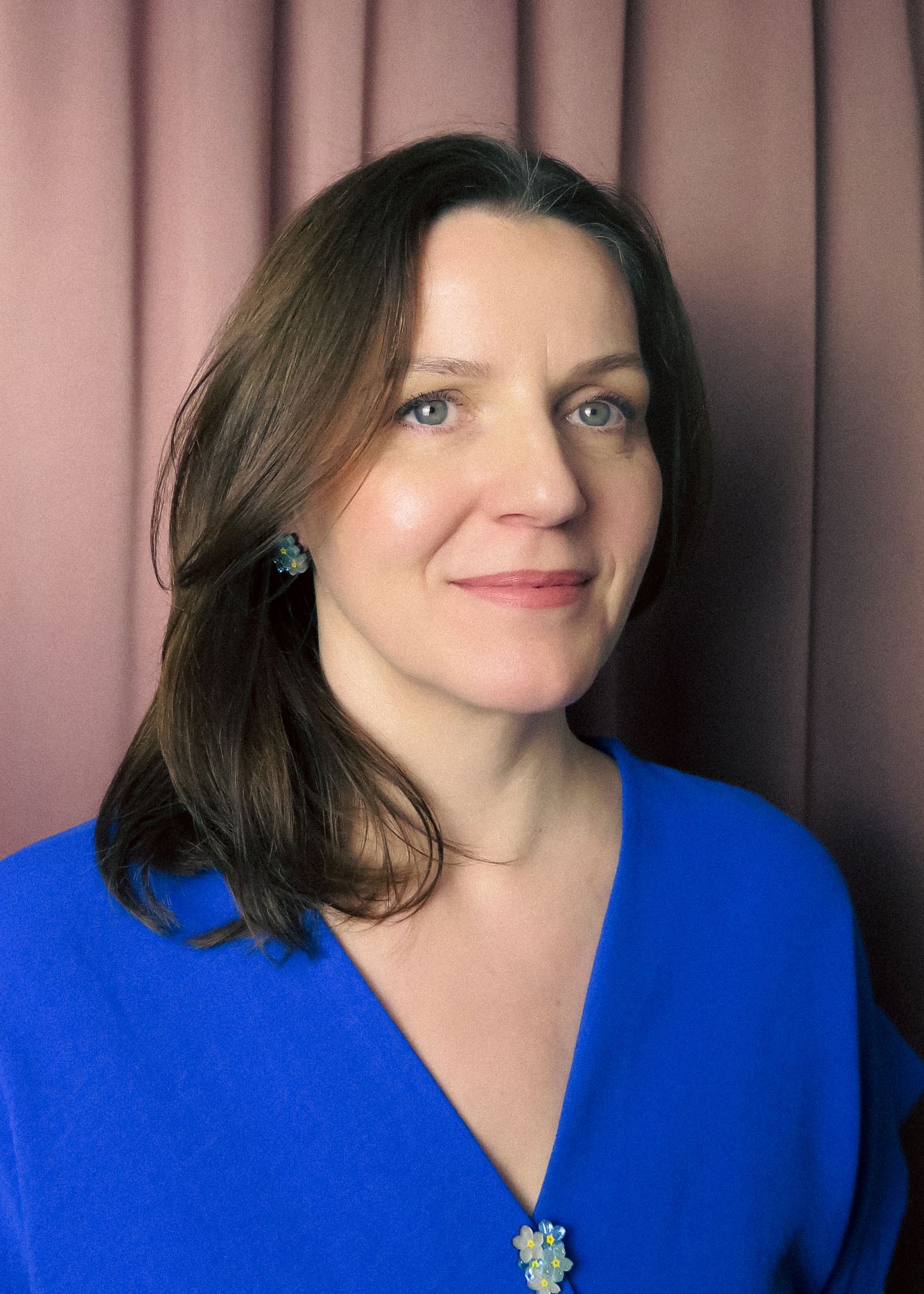 A woman with shoulder-length brown hair wearing a blue top and a handmade Forget Me Not Flower lapel pin stands in front of mauve curtains, smiling gently at the camera.