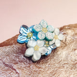 A cluster of delicate, handmade Forget Me Not Flower lapel pins with pastel blue and white petals and yellow centers rests on a textured rock against a soft pink background.