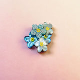 A cluster of handmade blue and white Forget Me Not Flower lapel pins with yellow centers is arranged on a smooth, pastel pink and peach gradient background.