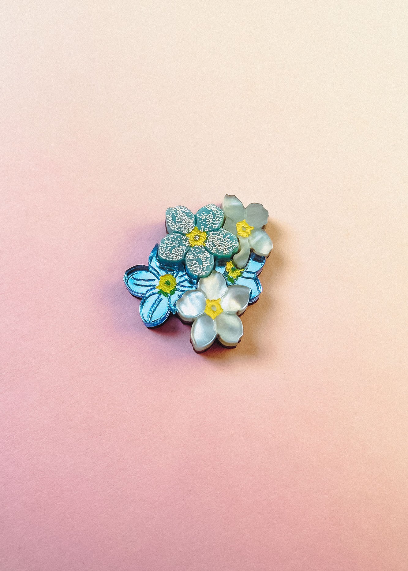 A cluster of handmade blue and white Forget Me Not Flower lapel pins with yellow centers is arranged on a smooth, pastel pink and peach gradient background.