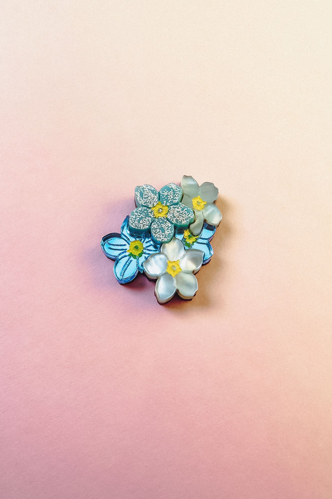 A cluster of handmade blue and white Forget Me Not Flower lapel pins with yellow centers is arranged on a smooth, pastel pink and peach gradient background.