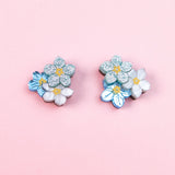 A pair of floral earrings, the Forget Me Not Studs, featuring blue, white, and glittery silver flowers are displayed on a light pink background—wearable art that brightens any look.