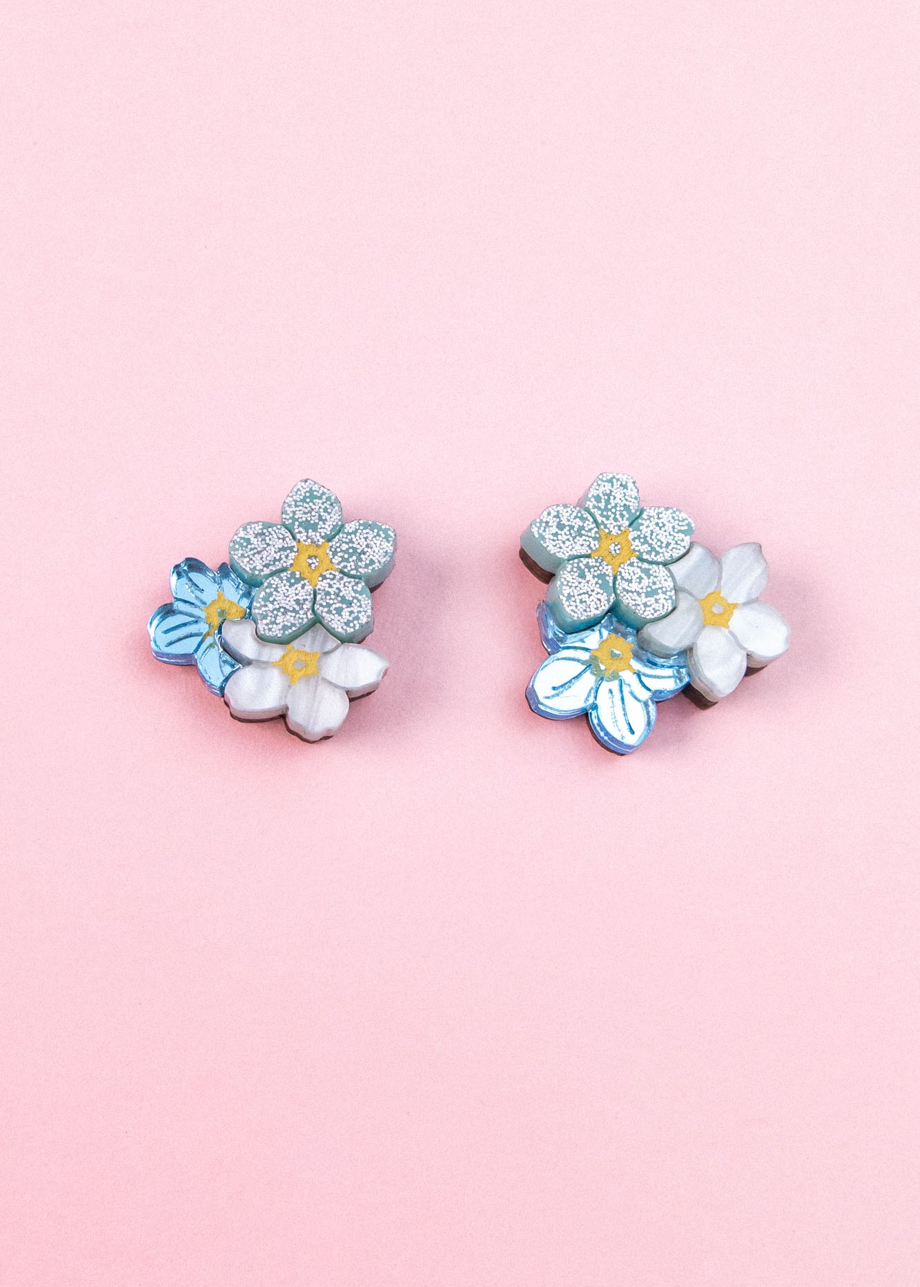 A pair of floral earrings, the Forget Me Not Studs, featuring blue, white, and glittery silver flowers are displayed on a light pink background—wearable art that brightens any look.