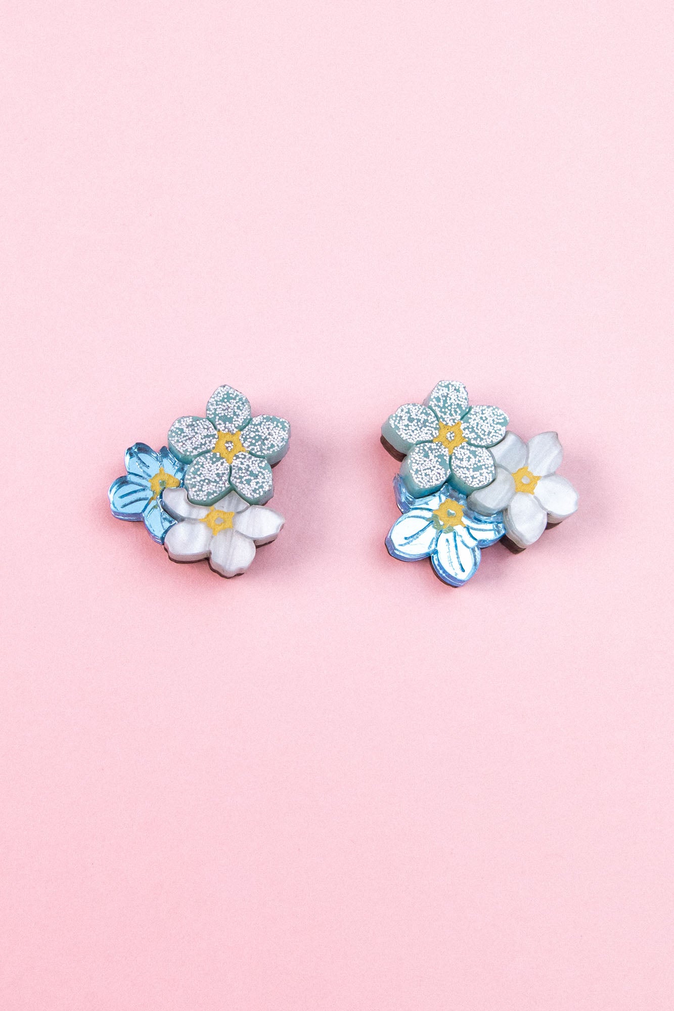 A pair of floral earrings, the Forget Me Not Studs, featuring blue, white, and glittery silver flowers are displayed on a light pink background—wearable art that brightens any look.