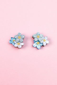 A pair of floral earrings, the Forget Me Not Studs, featuring blue, white, and glittery silver flowers are displayed on a light pink background—wearable art that brightens any look.