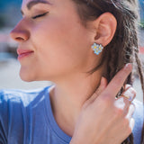 A woman with braided hair and Forget Me Not Studs smiles softly with her eyes closed, gently touching her hair. She wears a light blue shirt and stands outdoors in bright sunlight, showcasing the floral earrings as delicate wearable art.