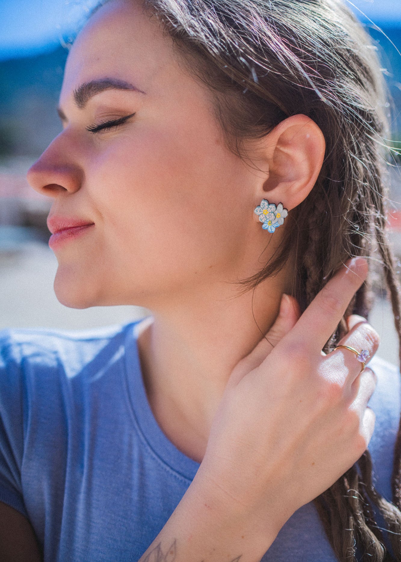 A woman with braided hair and Forget Me Not Studs smiles softly with her eyes closed, gently touching her hair. She wears a light blue shirt and stands outdoors in bright sunlight, showcasing the floral earrings as delicate wearable art.