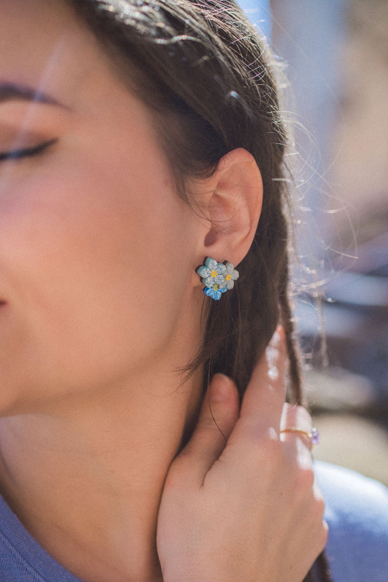 Close-up of a woman’s face, eyes closed, wearing Forget Me Not Studs—delicate floral earrings as wearable art. She touches her braided hair with one hand while sunlight softly illuminates her skin.