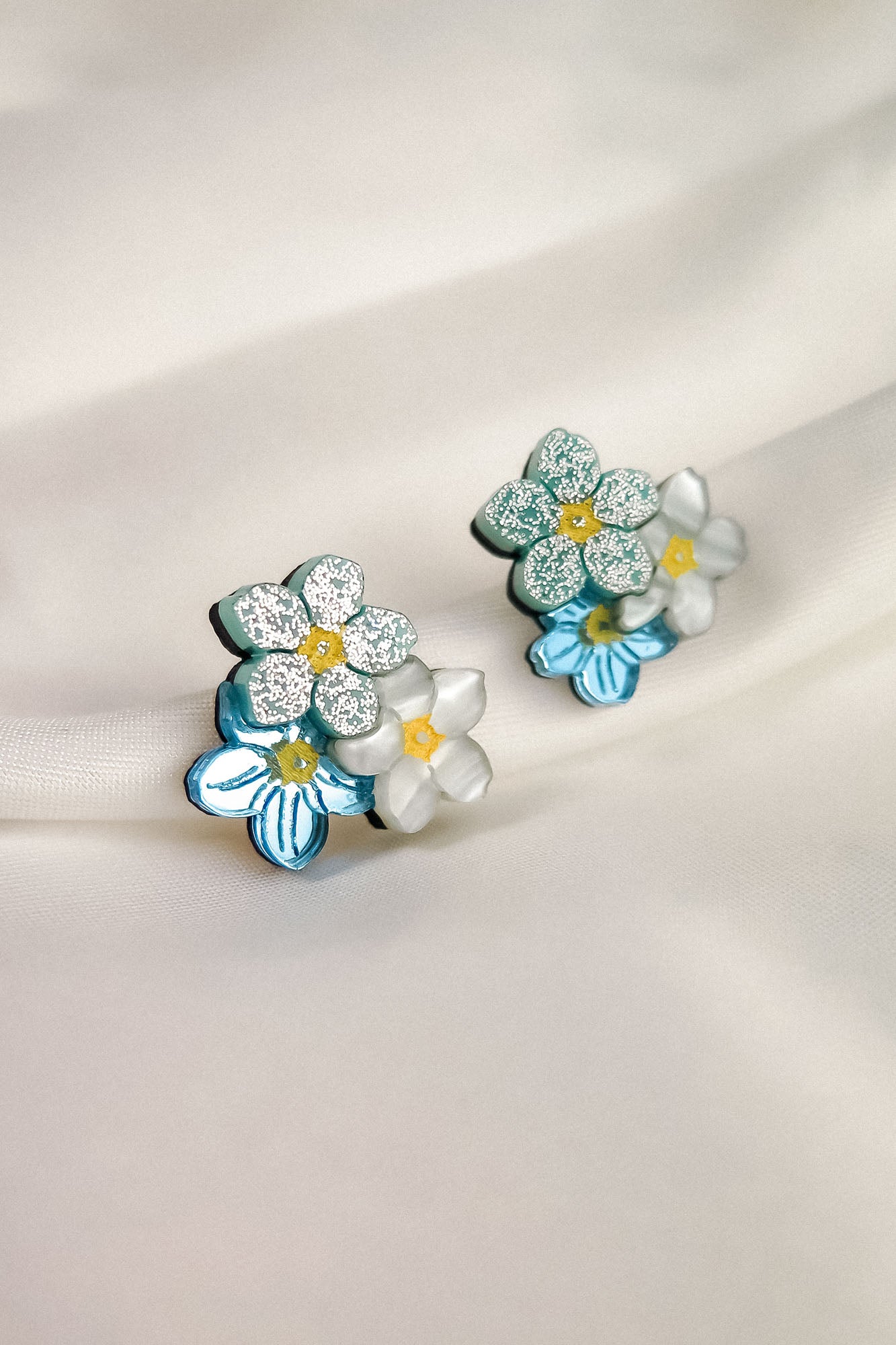 A pair of Forget Me Not Studs, featuring blue and white petals, yellow centers, and glittery accents, these floral earrings are true wearable art displayed on a soft, light-colored fabric background.