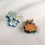 Two dainty floral earrings shaped like flowers rest on white fabric. One earring shows blue and white glitter details, while the other reveals the wooden back and metal post—these are the charming Forget Me Not Studs.