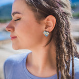 A woman with closed eyes and braided hair smiles softly outdoors, wearing a light blue shirt and Forget Me Not Studs—delicate floral earrings that are true wearable art—against a blurred, sunny background.