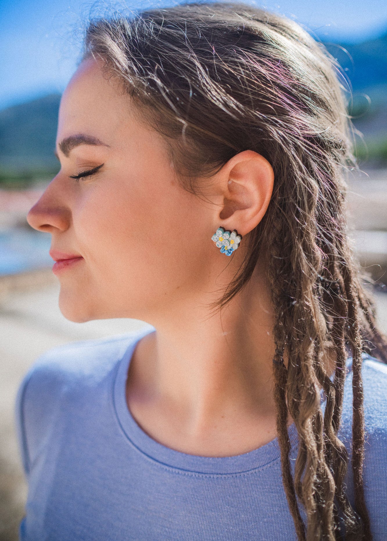 A woman with closed eyes and braided hair smiles softly outdoors, wearing a light blue shirt and Forget Me Not Studs—delicate floral earrings that are true wearable art—against a blurred, sunny background.