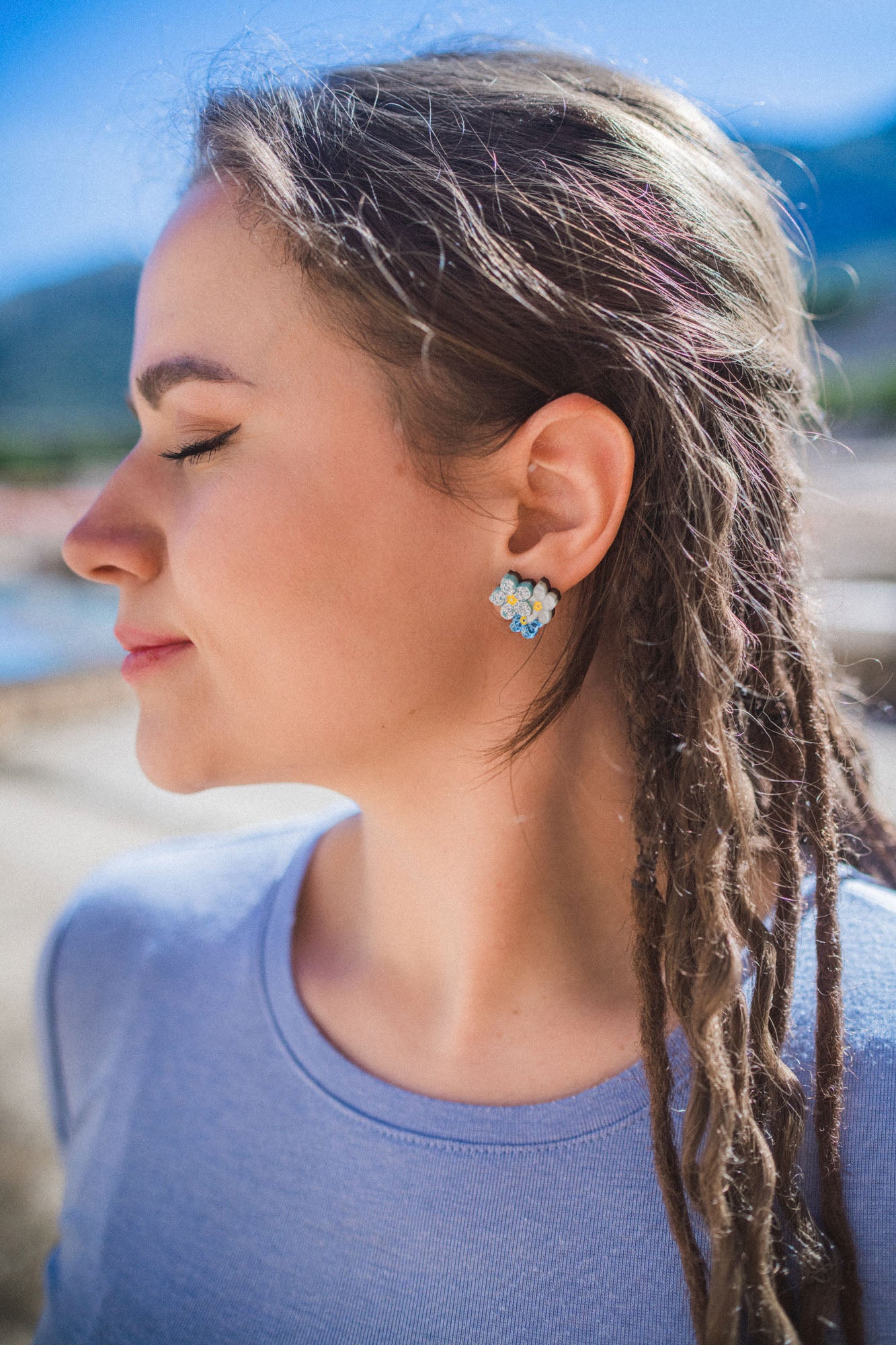 A woman with closed eyes and braided hair smiles softly outdoors, wearing a light blue shirt and Forget Me Not Studs—delicate floral earrings that are true wearable art—against a blurred, sunny background.