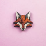 A geometric, stylized fox head brooch—handcrafted from recycled wood in shades of brown, black, and white—rests on a light pink background, making it an eco-conscious accessory with modern flair.