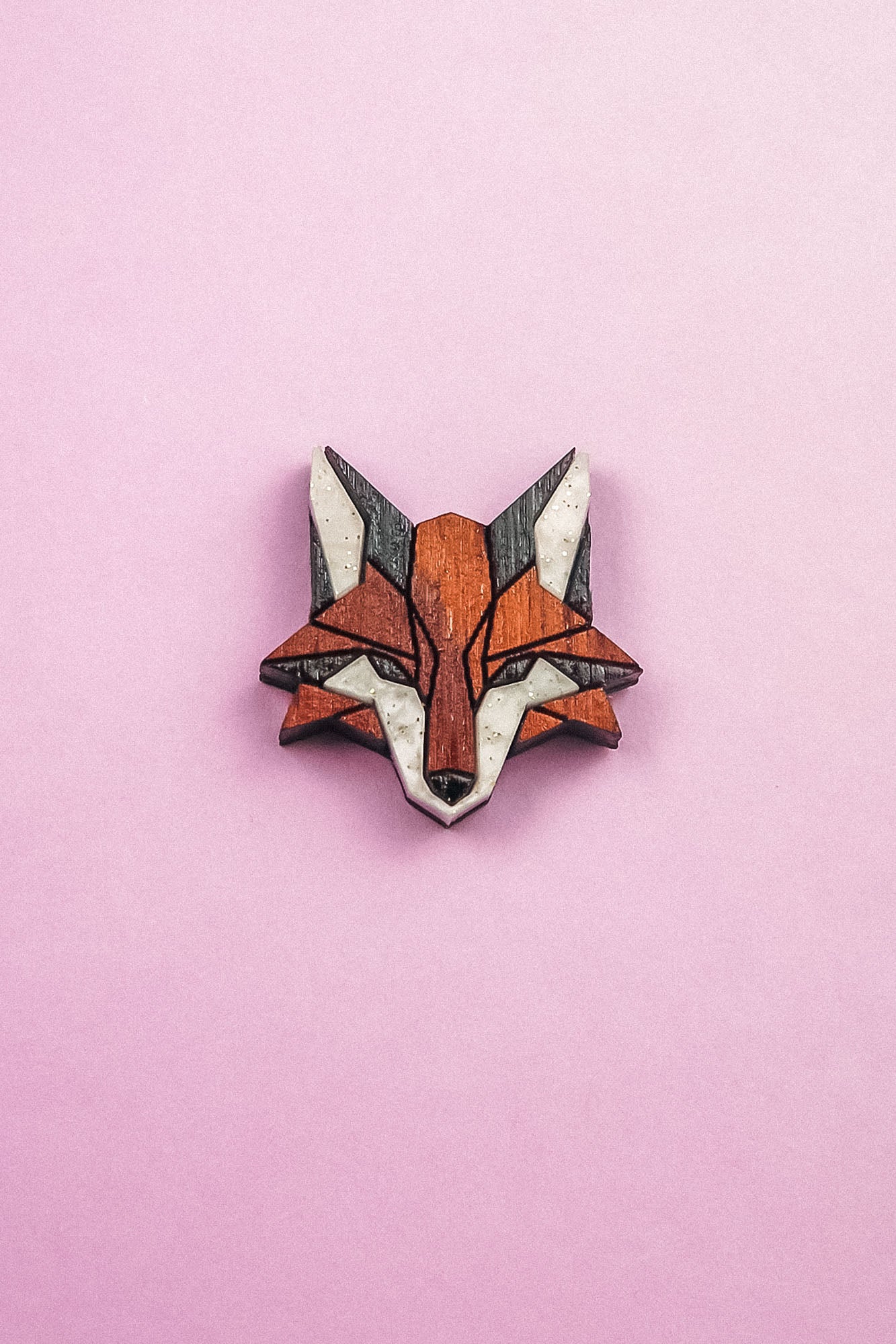 A geometric, stylized fox head brooch—handcrafted from recycled wood in shades of brown, black, and white—rests on a light pink background, making it an eco-conscious accessory with modern flair.