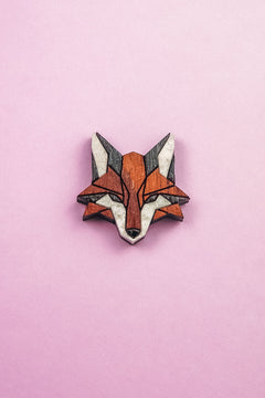 A geometric, stylized fox head brooch—handcrafted from recycled wood in shades of brown, black, and white—rests on a light pink background, making it an eco-conscious accessory with modern flair.