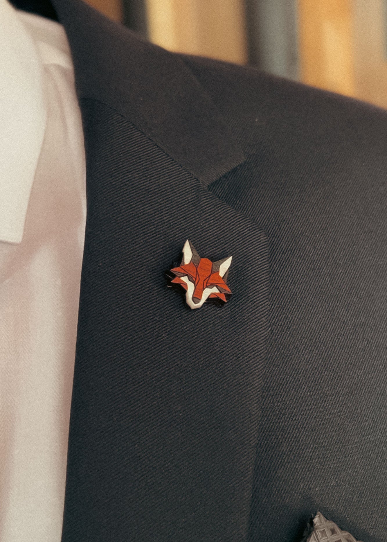 A close-up of a dark suit jacket with a handcrafted fox pin made from recycled wood, plus a hint of white dress shirt and patterned pocket square—an eco-conscious accessory for the modern gentleman.
