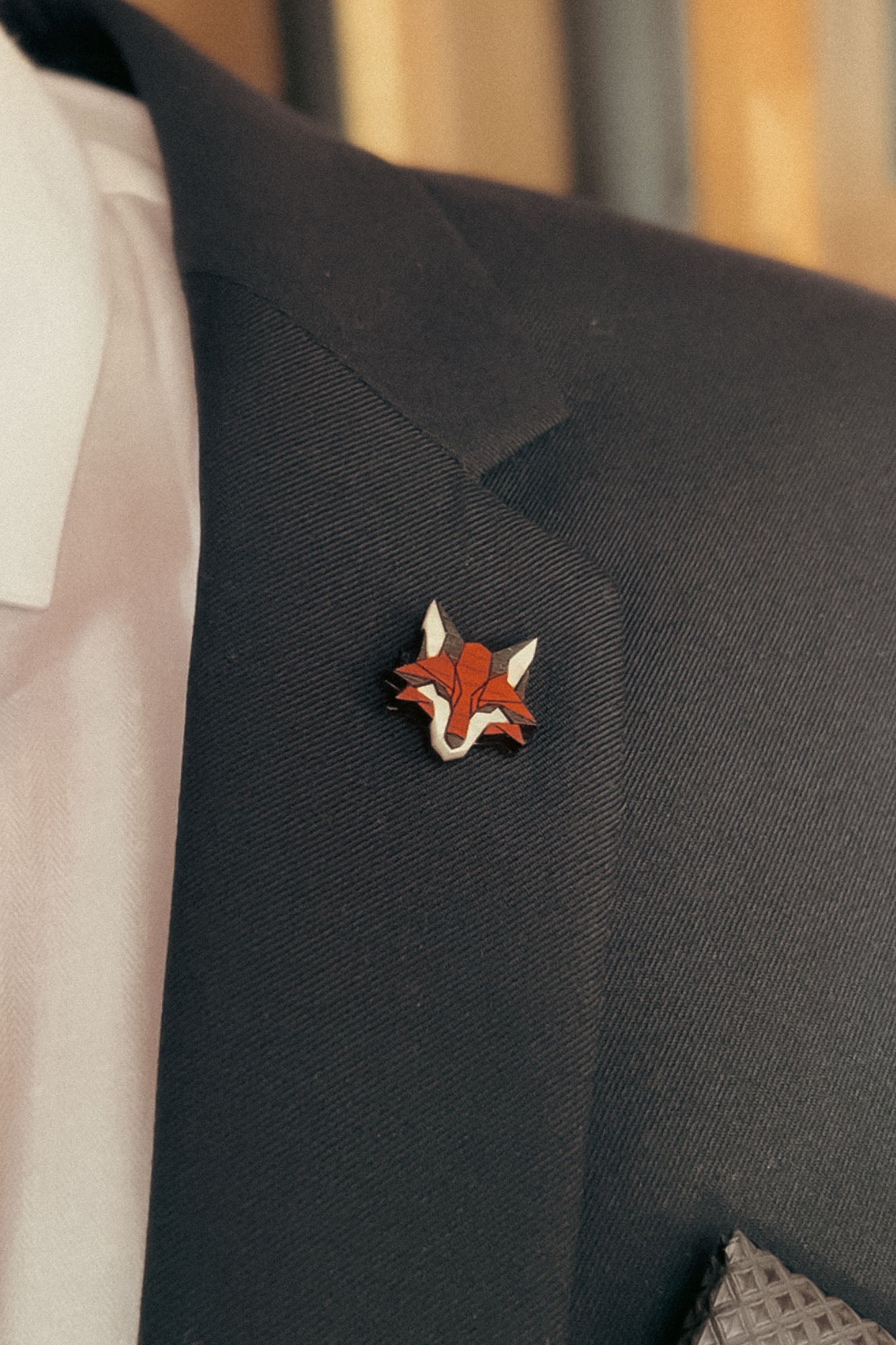 A close-up of a dark suit jacket with a handcrafted fox pin made from recycled wood, plus a hint of white dress shirt and patterned pocket square—an eco-conscious accessory for the modern gentleman.