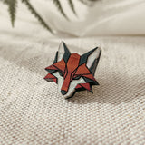 A geometric, handcrafted fox pin made from recycled wood rests on textured beige fabric, with blurred green fern leaves in the background.